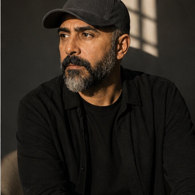 Arash seated at a table in warm window light, looking away from the camera.