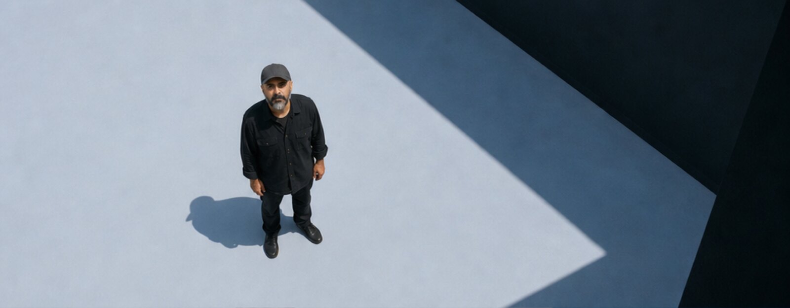 Overhead view of a lone figure in dark clothing standing on a pale mist-grey ground, crossed by a hard-edged diagonal shadow.